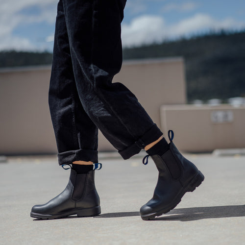 Person wearing black cuffed pants and black leather ankle boots walking outdoors on a concrete surface, with a blurred background of a building and distant trees under a blue sky.