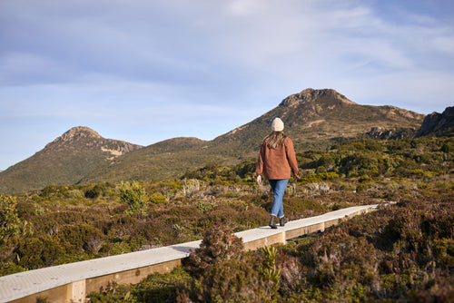 A person wearing a brown jacket and beanie walks along a wooden boardwalk through grassy, shrub-filled terrain with green hills and mountains in the background under a partly cloudy sky.