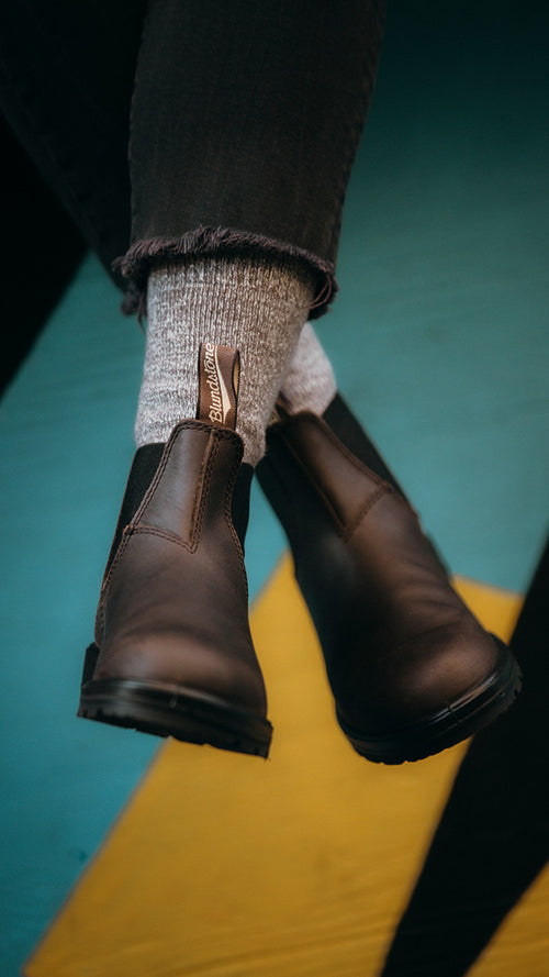 Close-up of a person wearing gray socks and dark brown Blundstone Chelsea boots, with frayed-hem black jeans, sitting against a blue and yellow background.