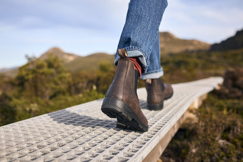 A person wearing brown boots and blue jeans walks on a raised metal pathway through a natural landscape with blurred hills and greenery in the background.