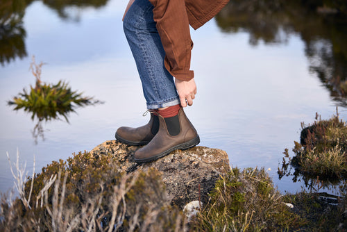 A person wearing brown boots, red socks, jeans, and a brown jacket stands on a rock near water, surrounded by greenery. The person is reaching down to adjust their pants.