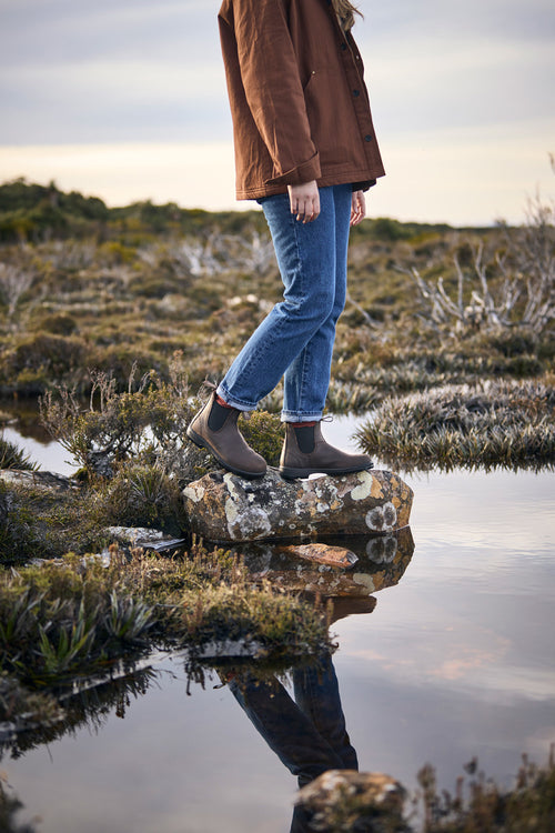 A person wearing jeans, brown boots, and a brown jacket stands on a rock surrounded by water and shrubs, with their reflection visible in the still water.