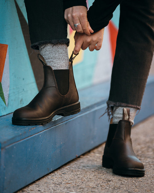 A person wearing gray socks and dark brown Chelsea boots pulls on a boot loop. They are standing on a sidewalk next to a blue wall with colorful geometric graffiti.