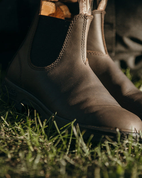 A close-up of brown leather Chelsea boots sitting on green grass in sunlight.