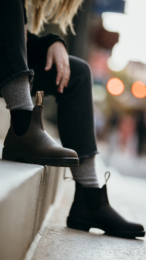 Person sitting on a curb, wearing dark brown Chelsea boots, gray socks, and black jeans. The background is softly blurred with warm lights and hints of an urban street scene.