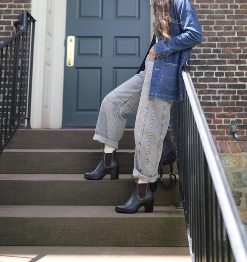 A person with long wavy hair sits on outdoor steps, dressed in a denim jacket, cuffed gray jeans, and black heeled boots. Only the lower face and body are visible. A dark door and brick wall are in the background.