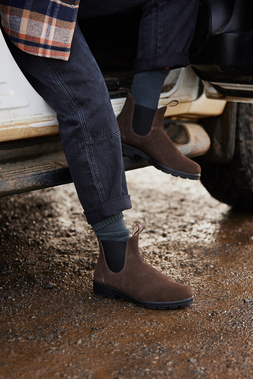 A person wearing dark jeans, green socks, and brown suede Chelsea boots steps out of a white vehicle onto a muddy ground. Only the lower legs and feet are visible.
