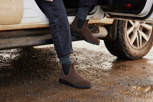 A person wearing dark jeans and brown suede Chelsea boots steps out of a muddy white vehicle onto wet, muddy ground. Only the lower legs and feet are visible.