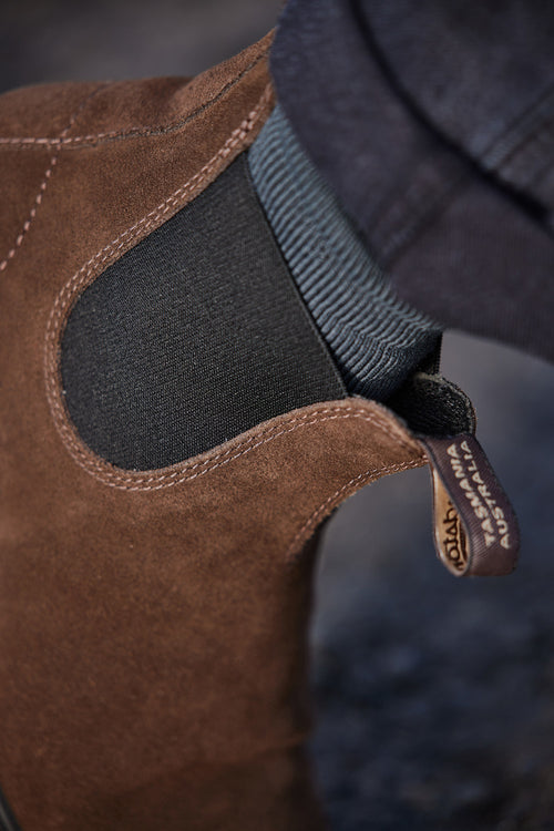 Close-up of a persons ankle wearing a black sock and a brown suede Chelsea boot with elastic side panel and a visible branded pull tab.