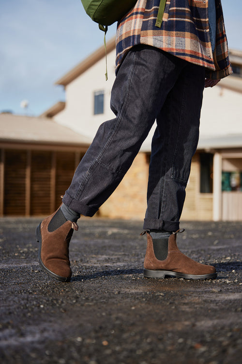 A person wearing brown Chelsea boots, cuffed dark jeans, and a plaid shirt stands on a gravel surface outside, with a blurred house and wooden structure in the background.