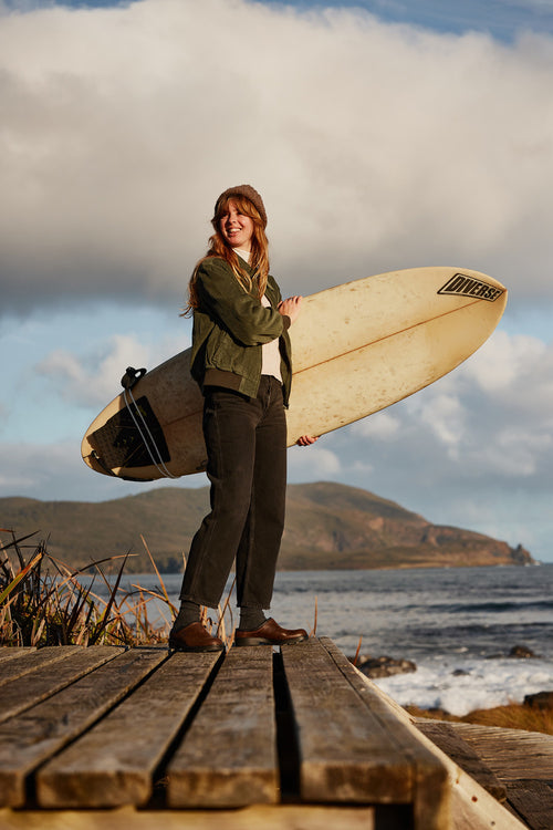 A young woman stands on a wooden boardwalk by the ocean, holding a surfboard under her arm. She is smiling, dressed casually in a jacket, jeans, and boots. The sky is partly cloudy and hills are visible in the background.