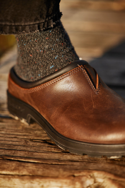 A close-up of a persons foot wearing a brown leather clog and speckled dark socks, standing on weathered wooden boards.