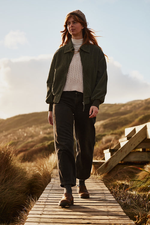 A woman with long hair walks confidently on a wooden path outdoors, wearing a green jacket, white sweater, dark pants, and brown shoes. The background features grassy hills and a partly cloudy sky.