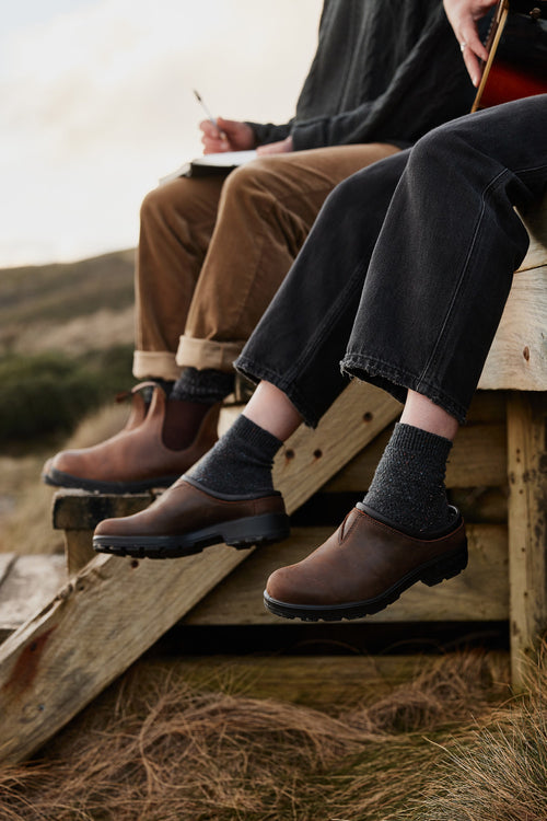 Two people sit on wooden steps outdoors, wearing dark socks and brown slip-on shoes. One person holds a notebook, the other holds a guitar. Grass and natural landscape are visible in the background.