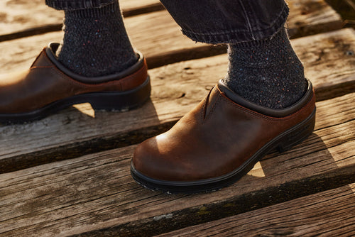 Close-up of a person wearing dark speckled socks and brown leather slip-on shoes, standing on weathered wooden planks outdoors.