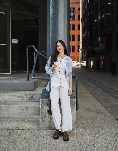 A woman with long dark hair, dressed in a light blue shirt over a white blouse and white pants, stands outdoors leaning against a pole, holding a coffee cup and looking relaxed on a city street.
