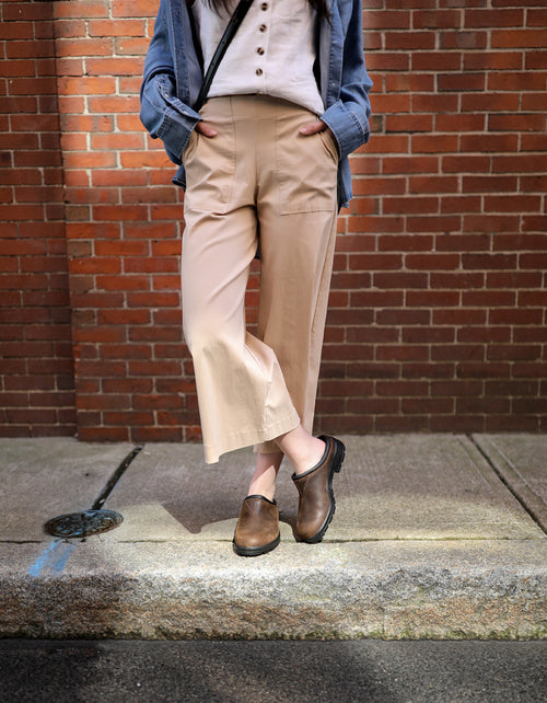 Woman standing on a sidewalk against a brick wall, wearing beige wide-leg pants, a white shirt with buttons, a blue overshirt, and brown clogs. Her hands are in her pockets and her face is not visible.