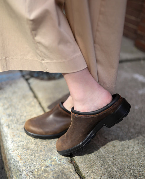 A person wearing beige pants and brown leather clogs stands on a concrete curb with one heel slightly lifted, exposing the back of their foot.