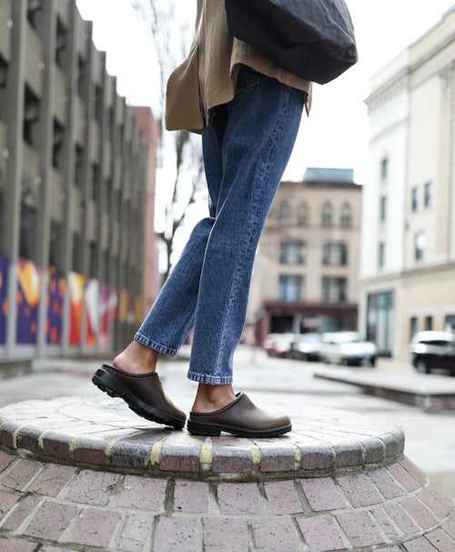 Person wearing blue jeans and brown clogs stands on a brick circular platform in an urban setting with modern and historic buildings in the background.