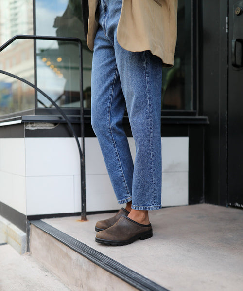 A person wearing blue jeans, brown slip-on shoes, and a beige coat stands on a concrete step outside a building with glass windows and a black metal railing.