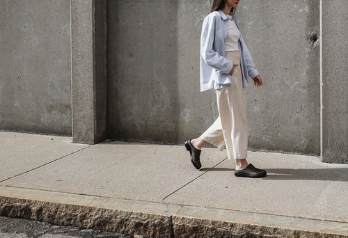 A person wearing a light blue shirt, white top, and cream pants walks along a concrete sidewalk next to a gray wall.