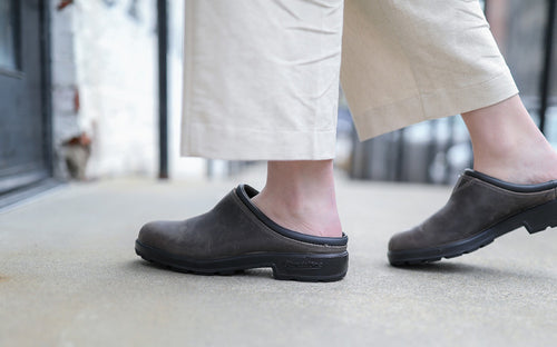 A person wearing light-colored pants and dark slip-on clogs is walking on a concrete sidewalk. The photo is focused on their legs and feet.