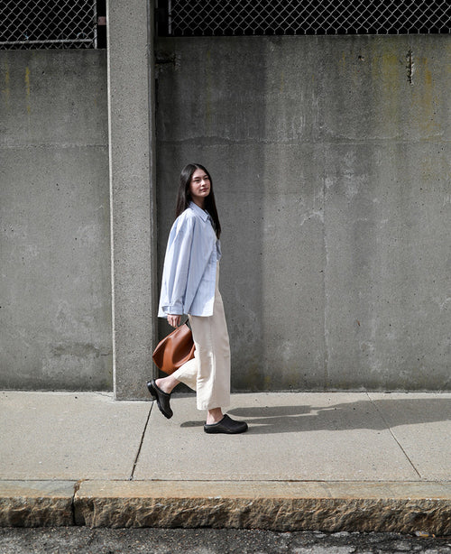 A woman with long dark hair walks on a sidewalk beside a concrete wall, wearing a light blue oversized shirt, beige pants, black shoes, and carrying a brown bag.