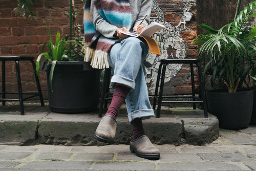 A person wearing cuffed jeans, brown boots, and a colorful scarf sits on a stone ledge outside, writing in a notebook, surrounded by potted plants and a brick wall with a mural.