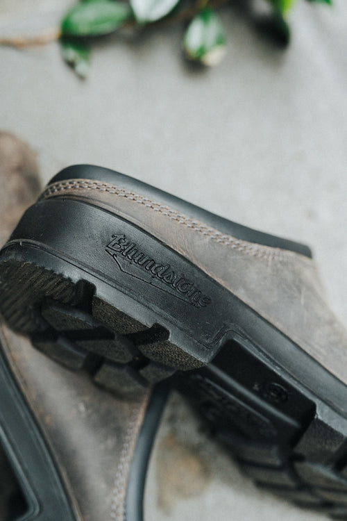 A close-up of a rugged, brown shoe with black treaded soles. The word Blundstone is embossed on the side of the sole, and there are green leaves blurred in the background.