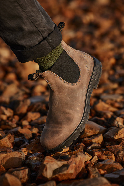 A close-up of a person wearing a brown leather Chelsea boot with green socks and black pants, standing on a ground covered with reddish-brown rocks.