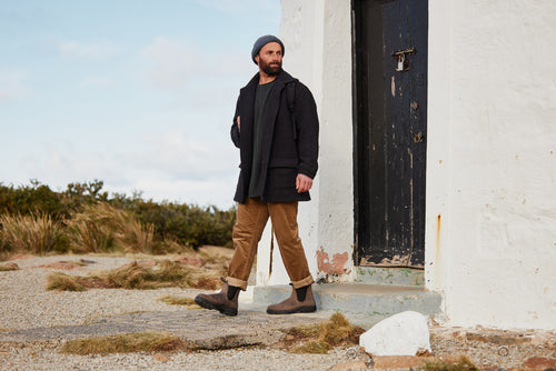 A man wearing a gray beanie, dark coat, scarf, brown pants, and boots walks outside next to a white building with a worn black door. The ground is rocky and some grass is visible in the background.