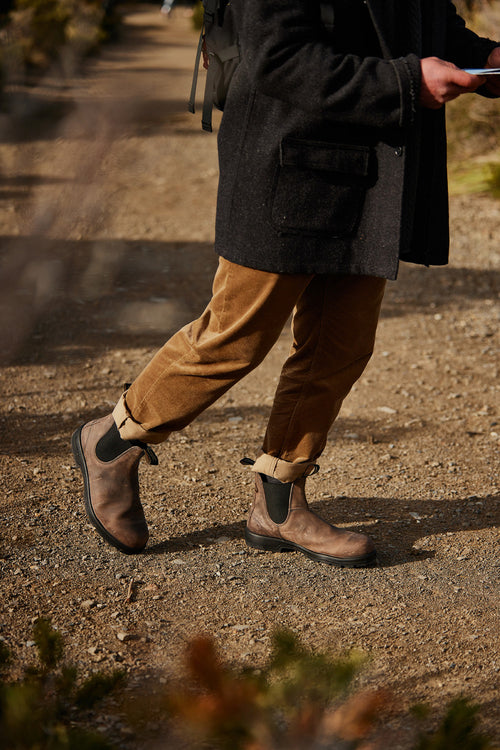 A person wearing rolled-up brown pants, brown Chelsea boots, and a dark coat walks on a dirt path, holding an object in one hand. The upper part of the person’s body is not visible.