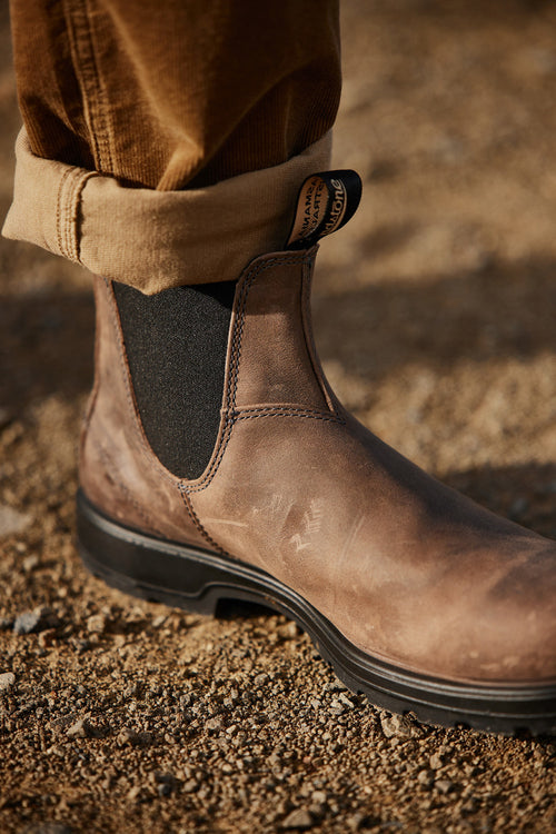 A close-up of a person wearing a brown leather Chelsea boot with black elastic side panels, standing on a gravel surface. The person’s tan pants are rolled up above the ankle.