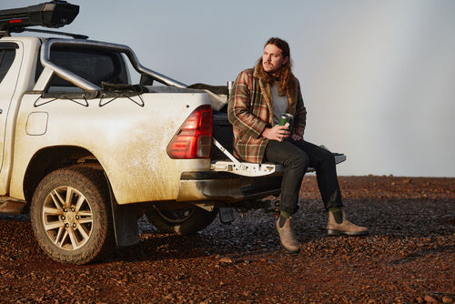 A man with long hair and a beard sits on the tailgate of a dirty white pickup truck, holding a cup. He wears boots, dark jeans, a green shirt, and a plaid jacket, looking off to the side on a rocky terrain.