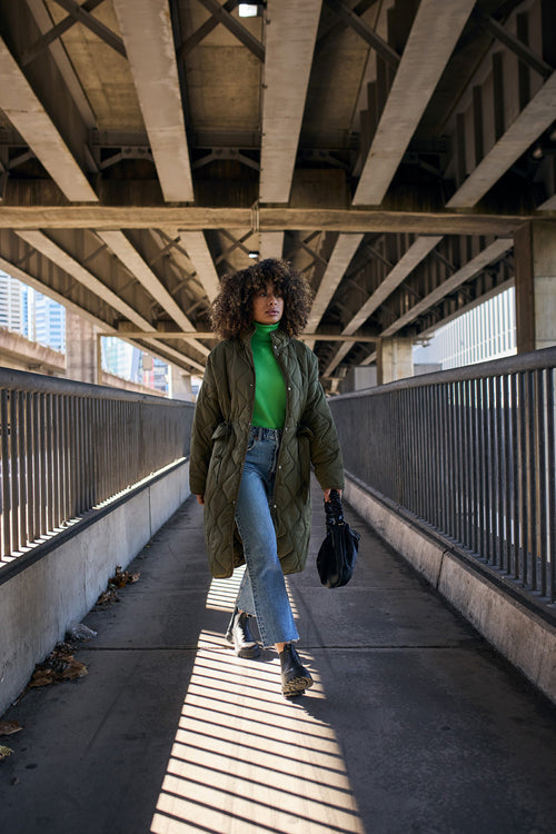 A person with curly hair walks confidently under a bridge, wearing a green turtleneck, long olive quilted coat, flared jeans, and boots, holding a black handbag. Sunlight creates shadows on the walkway.
