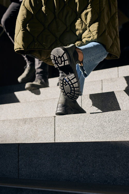 A close-up view of two people walking up stone steps, focusing on the sole of one persons black boot. They wear jeans and an olive green quilted coat in bright sunlight.