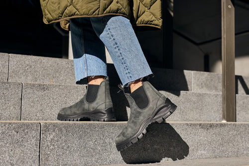 Close-up of a person wearing black Chelsea boots, cropped blue jeans, and a green quilted coat while walking up concrete steps in sunlight.