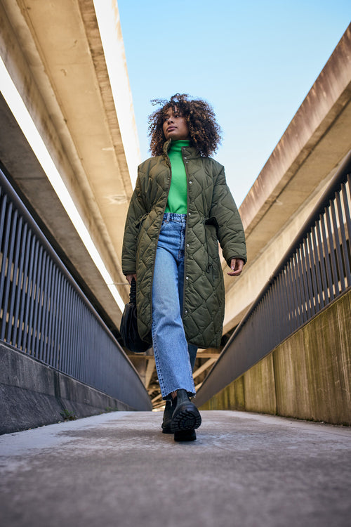 A woman in a green quilted coat, green turtleneck, blue jeans, and black boots walks confidently between two concrete walls under an overpass, holding a black bag. The sky is clear and blue above her.