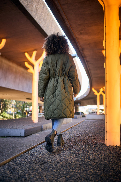 A person with curly hair, wearing a long quilted green coat and jeans, walks away under an overpass with glowing, tree-like lights on a gravel path.