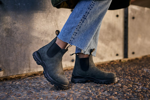 A person wearing rugged black Chelsea boots and frayed hem blue jeans stands on a gravel path next to a concrete wall.