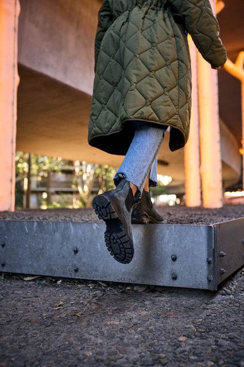 A person wearing a long, quilted green coat, cropped blue jeans, and black boots steps up onto a concrete platform outdoors, with orange-lit pillars in the background.