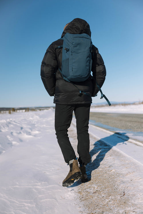 A person wearing a black winter jacket, dark pants, and boots walks on a snowy roadside with a blue backpack under a clear blue sky.