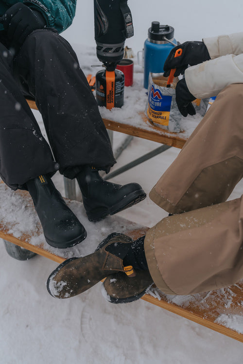 Two people in winter boots and gloves sit at a snowy wooden picnic table with camping gear, a blue thermos, and instant noodles, enjoying an outdoor meal in a snowy setting.