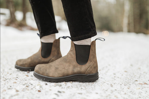Close-up of a person wearing brown suede Chelsea boots with black elastic panels, standing on a snowy, gravel-covered path. The person is also wearing light-colored socks and black pants.
