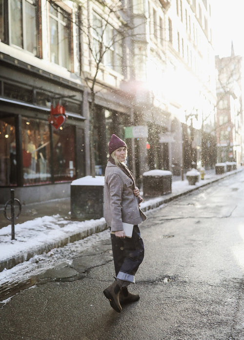 A person in a purple beanie and gray coat walks across a snowy city street in sunlight, turning to look back. There are snowbanks on the sidewalk and shop windows along the buildings.
