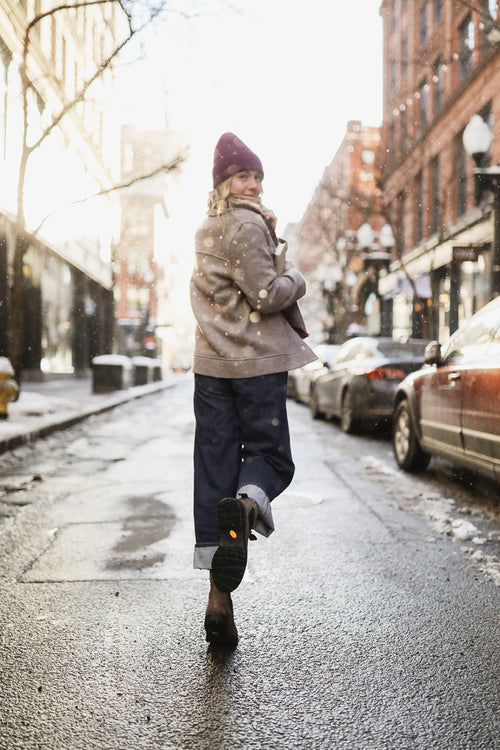 A person wearing a purple beanie, gray coat, and rolled-up jeans walks down a city street in winter, looking back and smiling. Snowflakes fall, and cars are parked along the sides of the wet road.