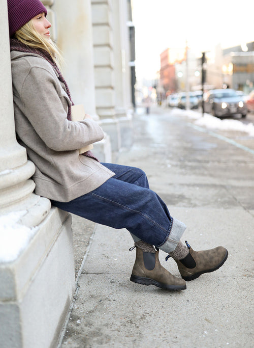 A person in a beige coat, blue cuffed jeans, brown boots, and a purple beanie sits against a building column on a snowy city sidewalk. Their head is turned away, and cars and buildings are visible in the background.