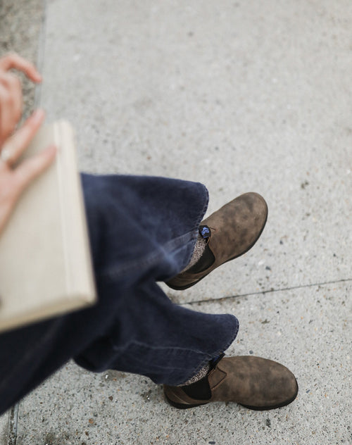 A person wearing dark blue jeans, brown shoes, and patterned socks sits on a light-colored concrete surface, holding a closed beige book in their left hand. The image is taken from above.