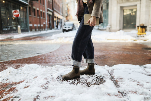 Person standing on a snow-covered street, wearing rolled-up jeans, brown boots, and holding a notebook. Only the lower half of the body is visible; brick buildings and snowy pavement are in the background.