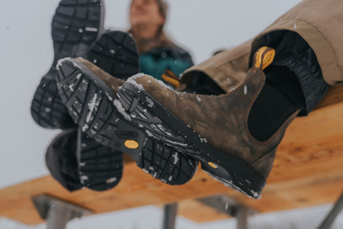 Close-up of two people sitting on a bench in winter, showing the soles of their boots dusted with snow. The background is slightly blurred, focusing on the boots and the snowy setting.